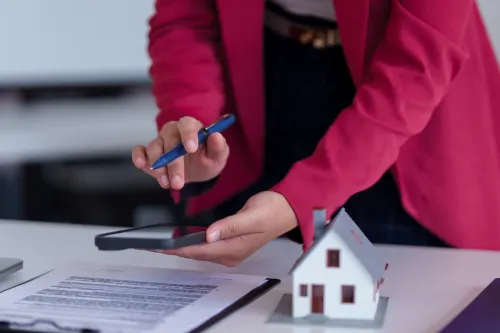 Profissional a verificar documentação com telemóvel, junto a uma casa em miniatura sobre a secretária